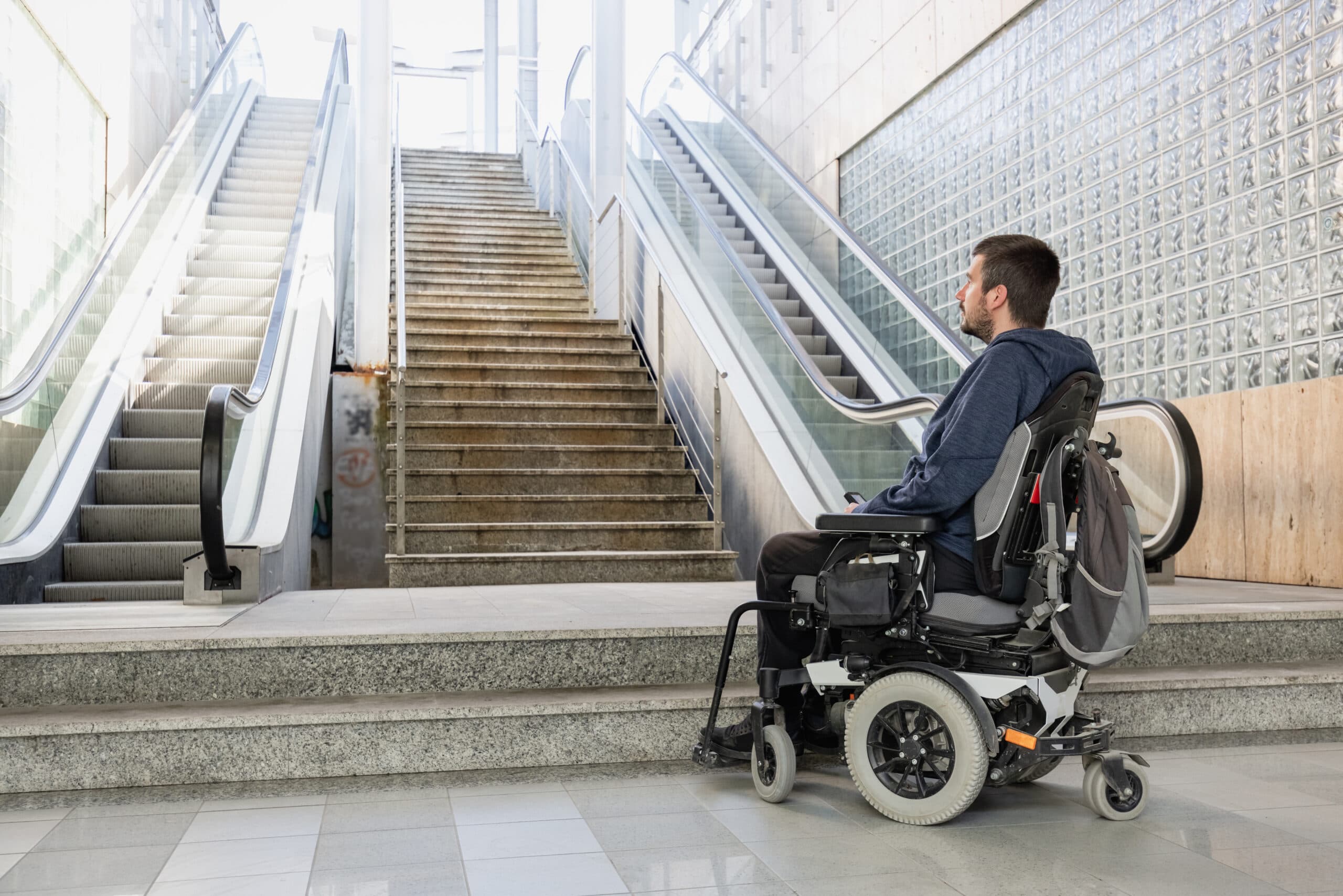 Un homme en fauteuil roulant face à un escalier flanqué de deux escaliers mécaniques