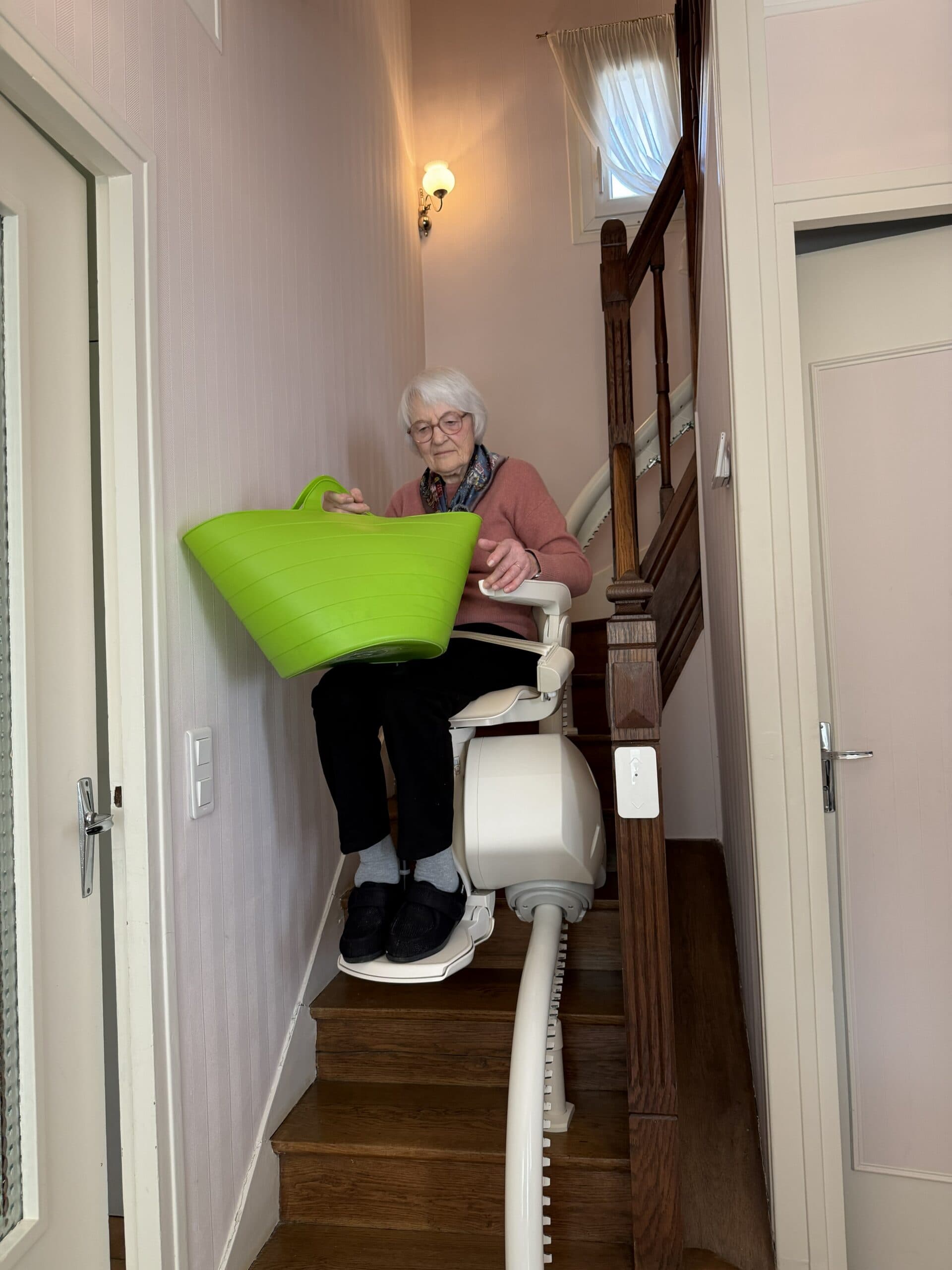 Une dame âgée assise sur son monte-escalier tenant un panier vert, à mi-parcours de l'escalier